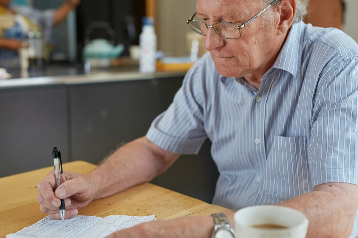 An older man in a striped shirt writes on paperwork at a wooden table with a cup of coffee beside him.